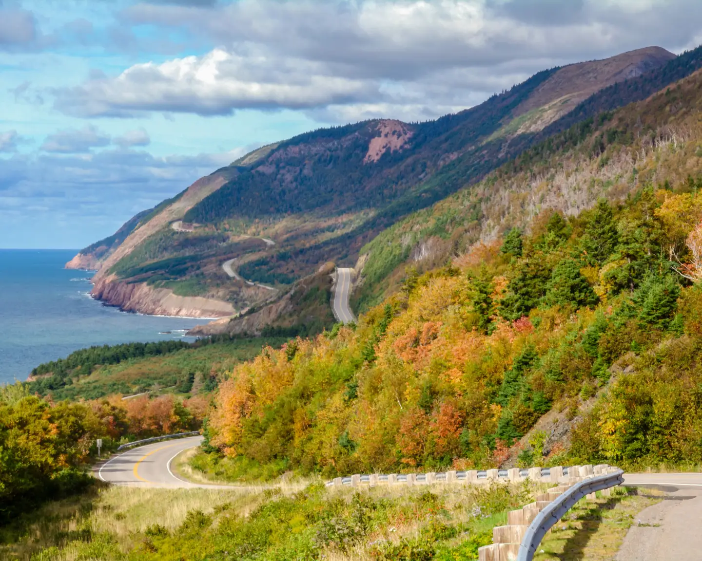Panoramablick auf die kurvenreiche Küstenstraße des Cabot Trail auf Cape Breton Island in Nova Scotia, Kanada, mit herbstlich gefärbten Wäldern, Bergen und dem Atlantik unter teils bewölktem Himmel