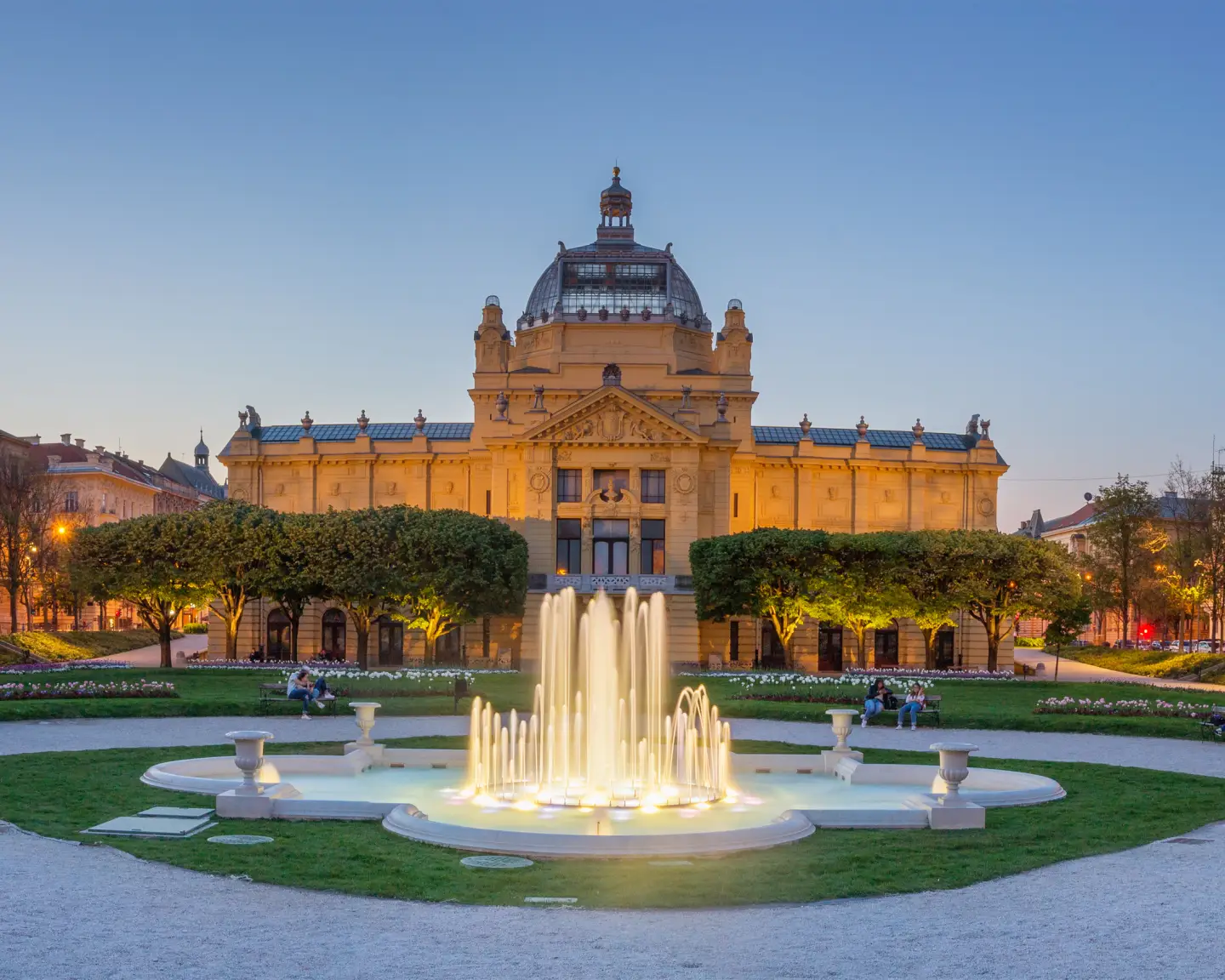 Pabellón de arte iluminado en Zagreb, Croacia, al atardecer, con una fuente en primer plano y un parque urbano bien cuidado rodeado de edificios históricos.