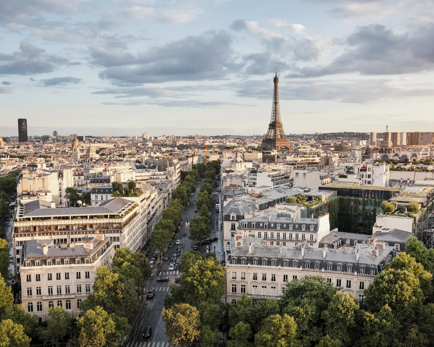 Panorama of Paris featuring the Eiffel Tower, wide tree-lined avenues, and classic Parisian architecture beneath a partly cloudy sky.