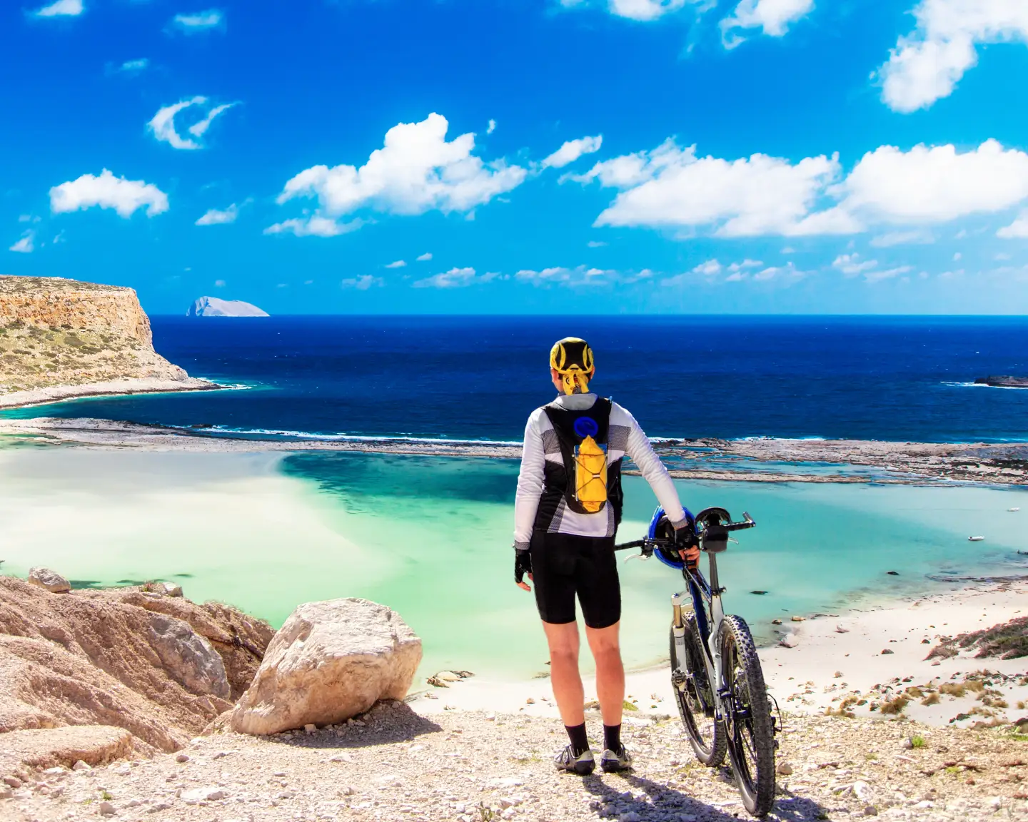 Een mountainbiker staat aan de oever van de Balos-lagune op Kreta, omringd door kristalhelder water en schilderachtige rotsformaties.