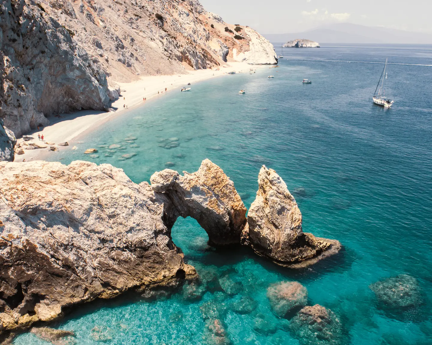 Kristallklares Meer an der Küste von Skiathos, Griechenland, mit markanten Felsen, natürlichem Felsbogen, weißem Sandstrand und Segelbooten unter strahlend blauem Himmel