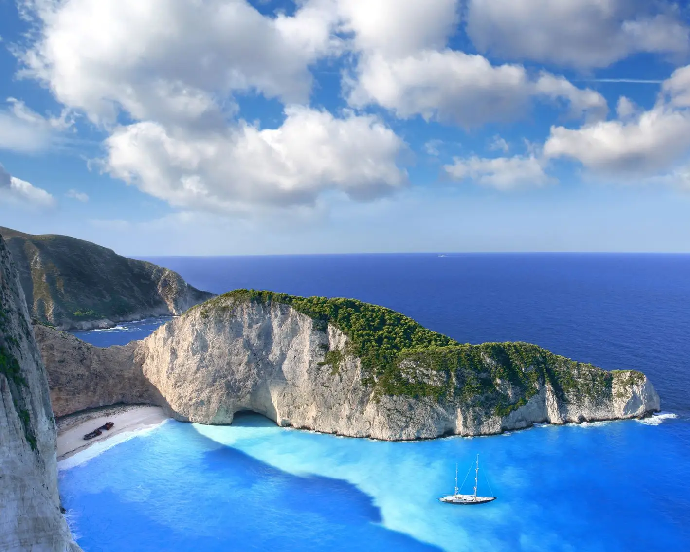 Vista panorámica de la famosa bahía de Navagio (bahía del naufragio) en Zante, Grecia, con aguas azul turquesa, escarpados acantilados blancos, un pecio en la playa y un velero en primer plano bajo un cielo ligeramente nublado.