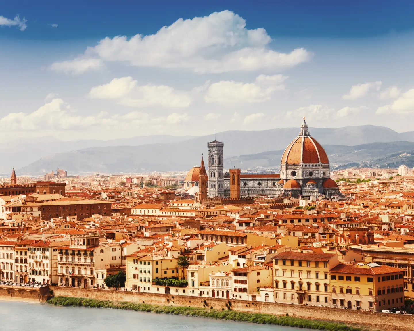 Panoramic view of Florence with its red roofs and cathedral. In the foreground, you can see the River Arno, with a range of hills in the background.