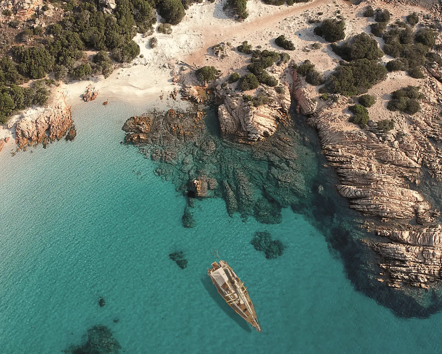 Vue aérienne d'une baie isolée en Sardaigne, avec des eaux turquoise claires, un rivage rocheux et un voilier à l'ancre.