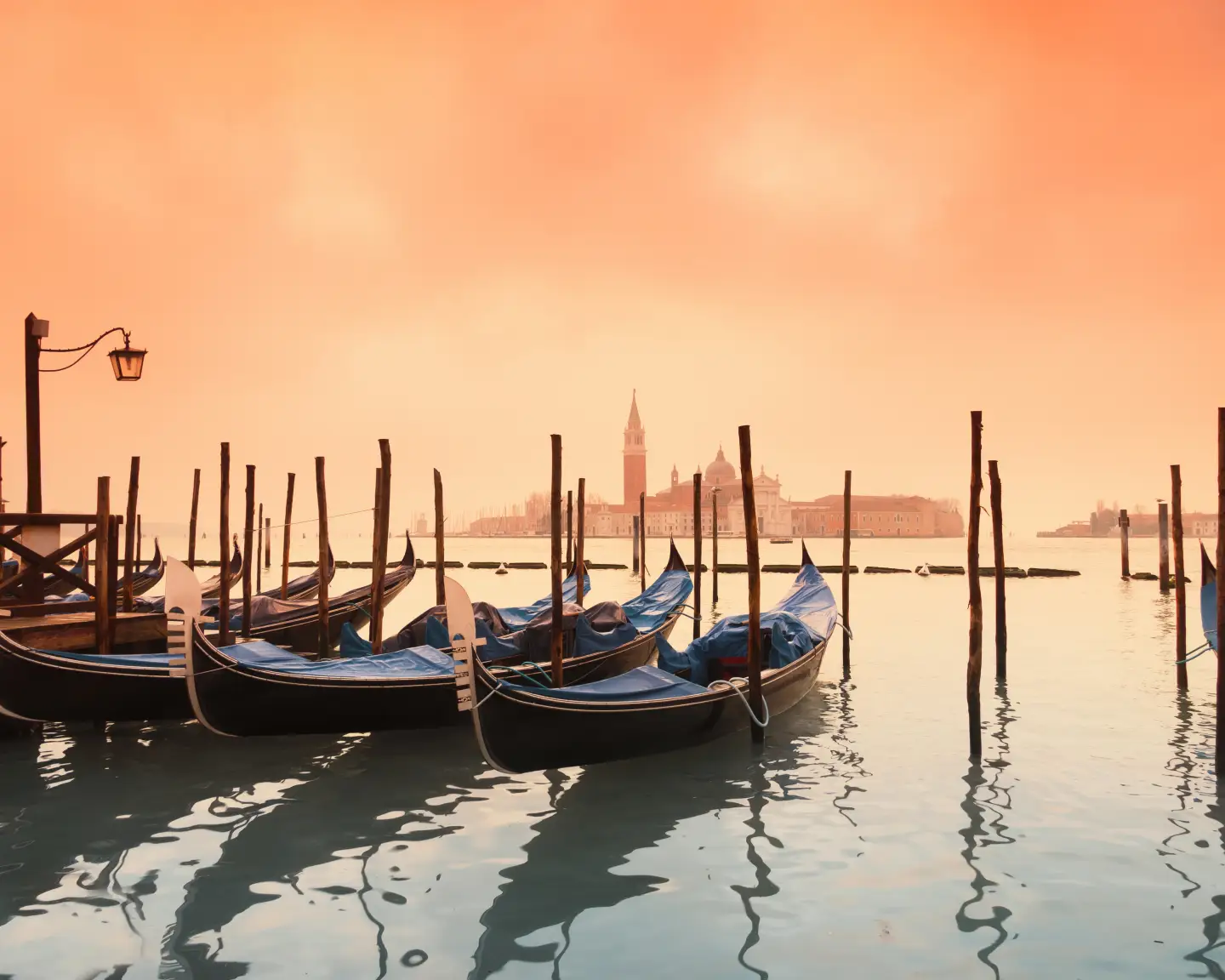 Calm waters reflect gondolas in the morning light, above which an orange sky stretches over the Basilica of San Giorgio Maggiore.