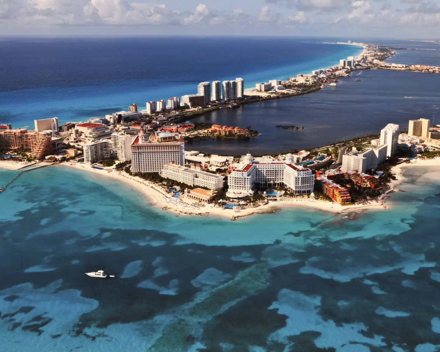 Vista aérea de la zona hotelera de Cancún, México, con playas de arena blanca, mar azul turquesa, grandes complejos hoteleros y la laguna de Nichupté bajo un cielo parcialmente nublado.