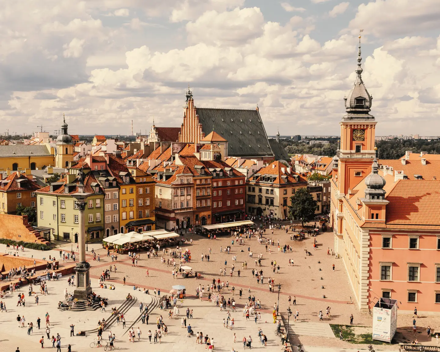 Vista aérea de la bulliciosa plaza del Castillo, en el casco antiguo de Varsovia, Polonia, con la columna de Segismundo, los vibrantes edificios históricos, el Castillo Real y la catedral de San Juan bajo un cielo parcialmente nublado.