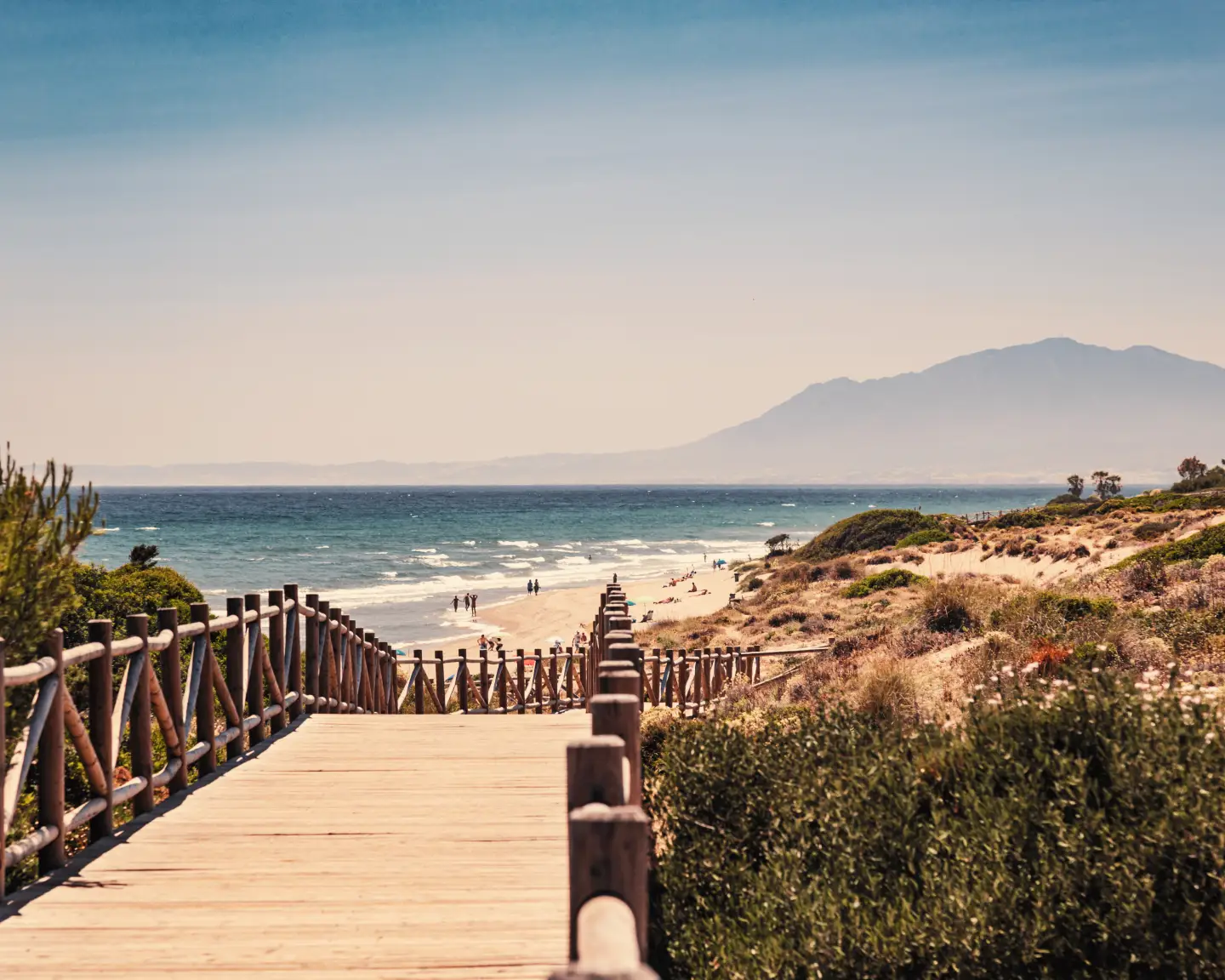 A beach in Málaga, Spain, with a wooden walkway leading to the sandy beach and the sea and mountains in the background, beneath a clear blue sky.