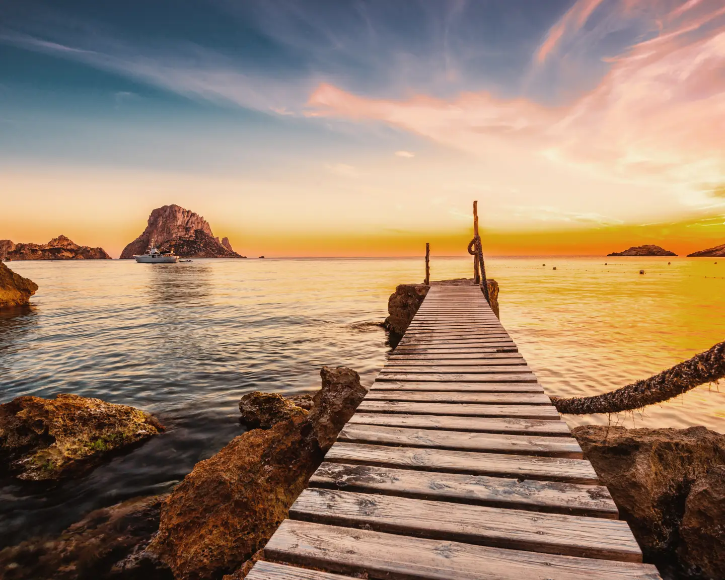 Sunset by the sea in Ibiza with a wooden jetty leading into the water and rocks in the background.