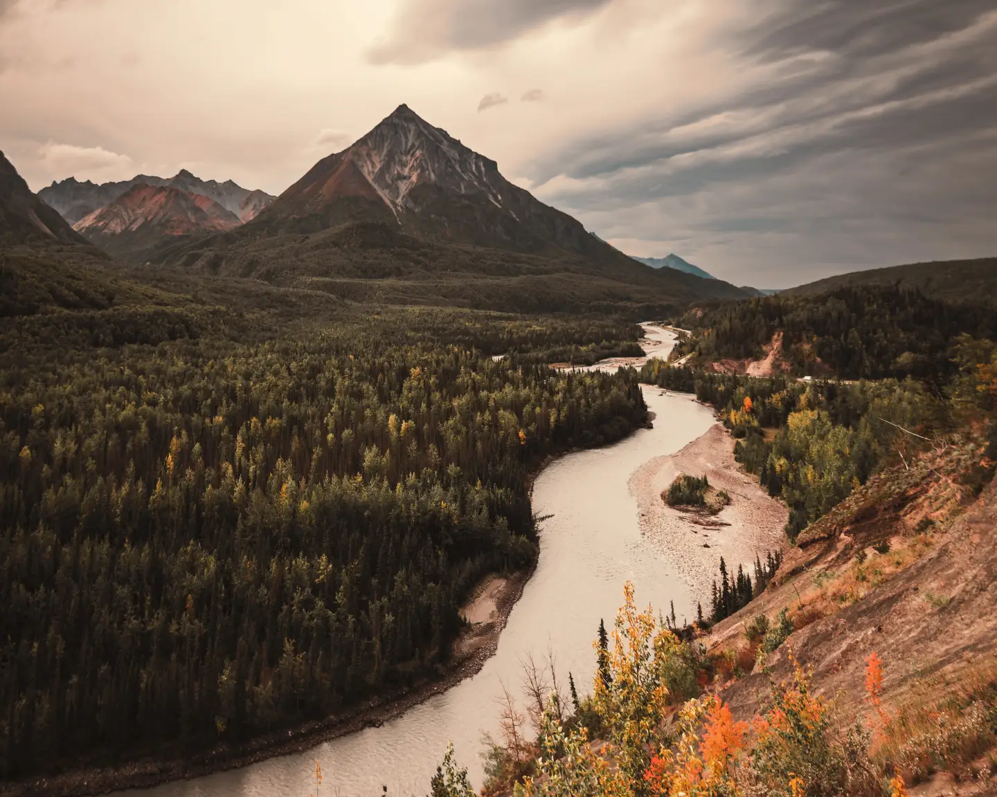 A picturesque river landscape with wooded hills and a high mountain in the background beneath a dramatic Alaskan sky.