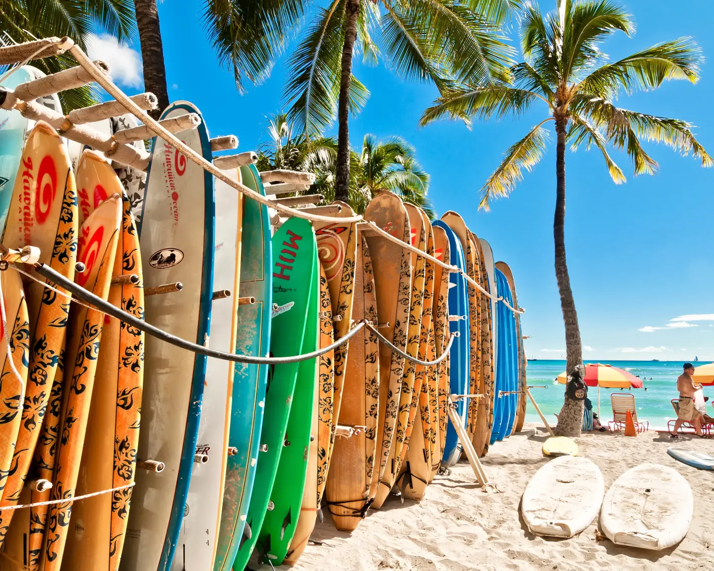 Farbenfrohe Surfbretter am sonnigen Strand von Honolulu unter Palmen, mit Blick auf das türkisfarbene Meer.